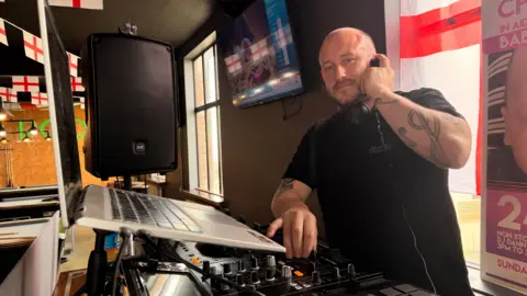 Jasmine Lowe/BBC News A man in a pub behind a DJ booth, he has decks and a laptop. One of his hands is spinning a deck the other is holding his headphones. He's looking at the camera standing next to a large speaker. He has blue eyes and tattoos. He looks fairly tired.