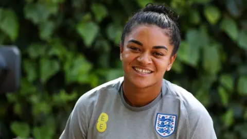 PA England international Jess Carter, whose black hair is tied up into a bun, wears a grey training shirt with the England badge on the front. She smiles for the camera.