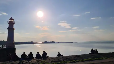Tom Richard A row of people sitting on a stone promenade beside a lighthouse, overlooking calm water as the bright sun hangs low in a clear sky.