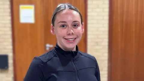 A woman in a black zip‑up athletic top stands indoors against light brick walls and wooden doors