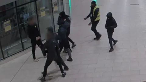 Teenagers. all clad in black with their faces covered, tussle in a shopping centre as a security guard moves in