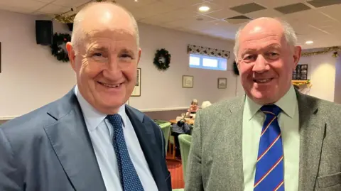 Vikki Irwin/BBC Kevin Hollinrake and Tim Passmore are standing together inside the Conservative club in Ipswich. You can see Christmas decorations and chairs and tables in the back ground. There is also a bright orange carpet in the background . Mr Hollinrake is on the left and is wearing a blue suit, pale blue shirt with a blue tie. Mr Passmore is wearing a green tweed suit, pale green shirt and blue red and yellow tie. Both men are smiling at the camera. 
