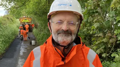 A man with a grey beard in an orange reflective jacket and white helmet with hedges behind him