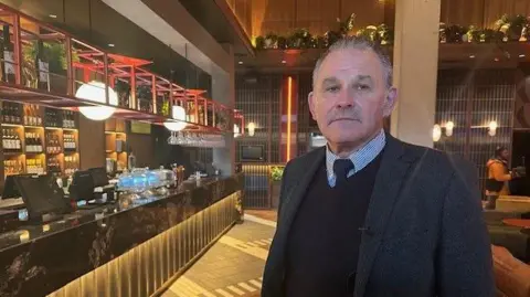 Allan Cook is suit and tie looking at camera in front of cinema bar with bright ball-shaped lights. 