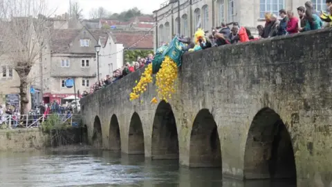Bradford-on-Avon Town Council Crowds of people stand on an old bridge in Bradford-on-Avon as bags of rubber ducks are thrown into the water. Near the bridge, people stand and watch.