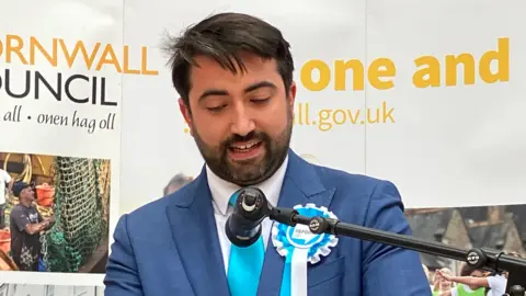 Bruce Craze seen speaking into a microphone while wearing a blue suit with a white shirt and light blue tie. He is also wearing a white and blue rosette with the Reform UK logo in the centre. Mr Craze has short, black hair and a short beard.