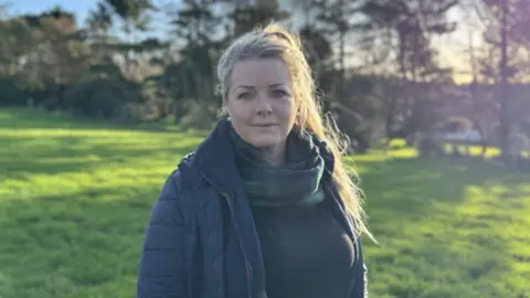 Kirree Kermode is standing in a green field lined with trees on a sunny morning. She has long blonde hair tied up and is wearing a navy coat with a green and blue tartan scarf.