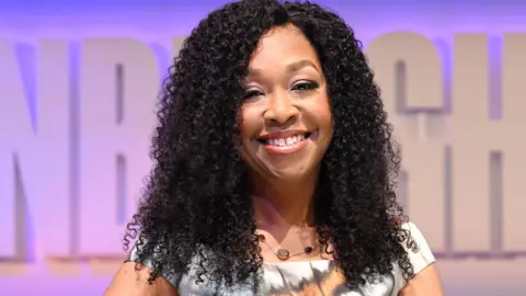 Shonda Rhimes, with a bright sile and dark curls, sitting on set at her TV festival show. The background is pale purple and she wears a patterned white, orange and black dress while sitting on a white chair.