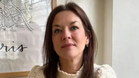 Lady with brown hair wearing a cream knit high neck jumper.