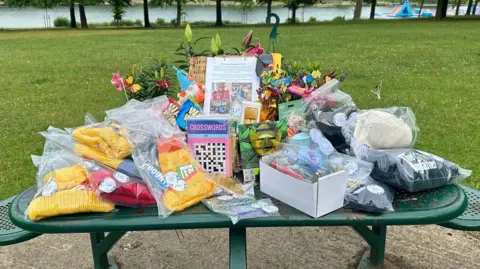Rachel Davies A green metal table is piled high with items. The table is in a park with a green lawn behind. On the table are items including knitted hats, puzzle books, flowers, garden ornaments and wrapped food.