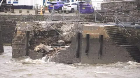 This picture shows a damaged harbour wall. A large section of the stone wall has collapsed, exposing rubble and debris inside. Water from the sea is flowing through the broken area, indicating structural failure likely caused by strong waves, erosion, or storm damage. There are metal safety barriers and fencing around the damaged section. In the background, there are parked cars and buildings. The water in the foreground looks rough.