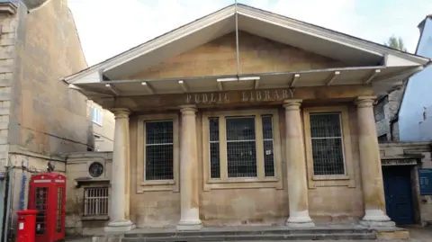 A sandstone fronted building with four columns, pitched roof and old glass windows. A sign on the front reads 'public library' and there is a red phone box and post box within shot.