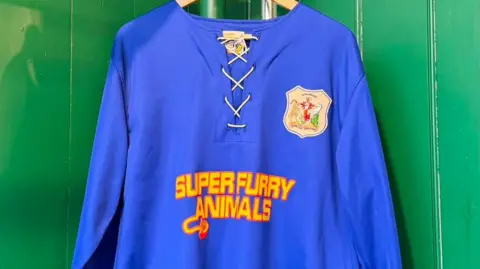 Eleri Williams Cardiff City and Super Furry Animals sponsored vintage shirt. It's a deep blue shirt with a vintage logo on the right hand side with yellow and orange writing saying 'Super Furry Animals'.  