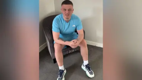 A man with short hair sitting on a chair wearing a blue top