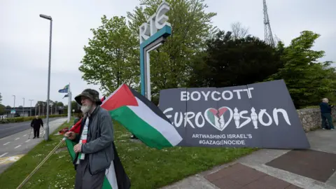 A man holding a Palestine flag walks past a sign which reads 'Boycott Eurovision.