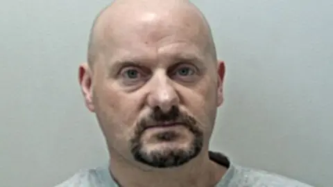 Anthony Croft's mugshot against a plain grey background. He has a bald head, brown goatee and is wearing a plain grey marl tshirt