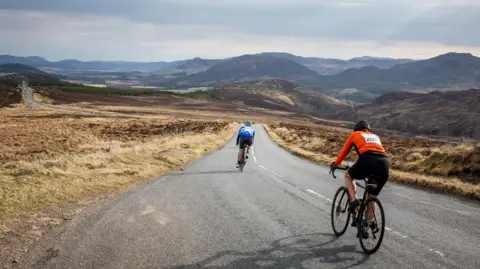 Paul Campbell Two cyclists, with their backs to the camera, free wheel down a hill on a long, rural road. Either side of the road is rugged moorland and low hills are in the distances. It is a fine day and the sun is out.