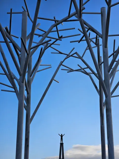Wade Campbell A statue of a person with their arms outstretched on top of a stand. Two tree-like sculptures are on each side. 