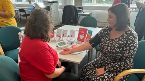 Two women sit at a table in a warm, community‑health setting. One, in a red shirt, is helping Sam Collier, who is wearing a patterned dress and has a blood‑pressure cuff on her arm
