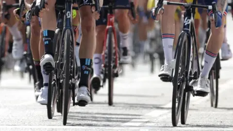 A large group of cyclists are riding along the road. They can only be seen from the legs down. 