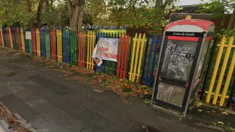 A phone box in front of a metal fence painted in alternating green, red, yellow and blue colours. 
