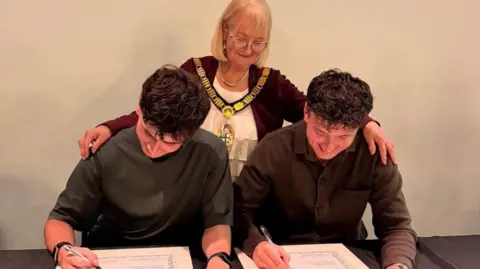 Redditch Borough Council The two brothers are sat at a desk signing certificates - behind them is the Mayor of Redditch, Sue Eacock, with a gold chain around her neck.
