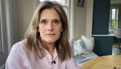 Ellen Roome is sitting on a sofa looking at the camera. She is wearing a pink and white pin-striped shirt and has medium-length brown hair and brown eyes.
