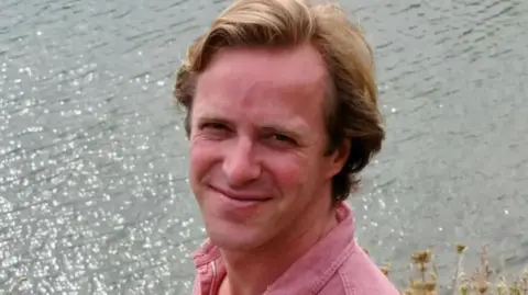 Thomas Kingston - a man in his 40s with dark blond hair - smiles as he stands near a lake. He is wearing a pink cotton shirt.