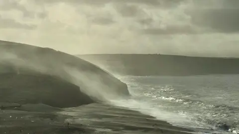 Jerry Betteridge Fine spray can be seen splashing up from the sea against rocks on the coast.
The land and sea appear grey with some glints of sun shining through.