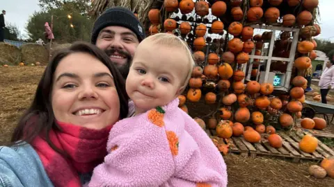 Gareth Williams A mum and dad in their thirties with a toddler in a pink fleece with pumpkins on it. It is around Halloween time with a display on pumpkins in the background in a muddy field. 