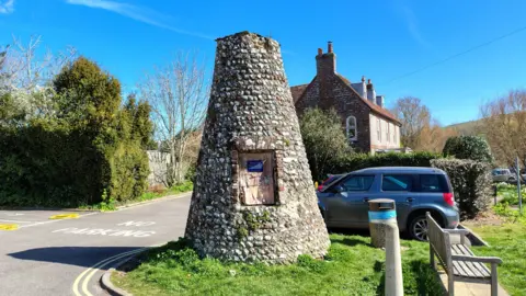 A flint stone obelisk by the side of a road. It is surrounded by a grass verge, with a car parked behind it, the road to the left of it, and a bench to the right of it.