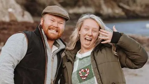 Laura with blonde hair and showing her ring and Ryan wearing a hat and stood next to her