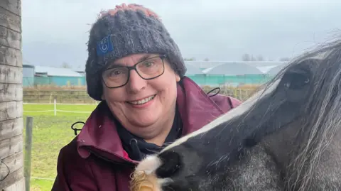 Mousumi Bakshi/BBC Alison Searle is on the left of the image. She is wearing a grey hat, burgundy coat and is wearing glasses. On the right is black and white horse whose head is in the image.
