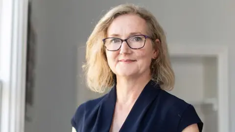 Resonance Photo of Rosie Phillips - CEO of Developing Health and Independence. She is looking at the camera, and is wearing a blue top, dark framed glasses and has blonde hair