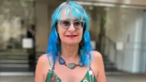 Sioux Vosper with blue hair wearing light grey glasses wearing a green patterned dress and a blue, green and orange shell patterned necklace. She is smiling. She is outside Dorland House in Paddington.
