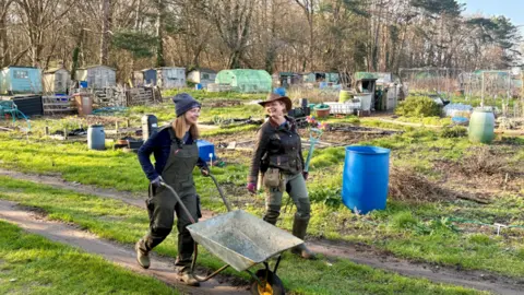 A woman holding a wheelbarrow in an allotment laughing with another woman carrying a gardening tool
