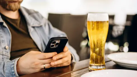 A man sitting in a pub looking at his mobile phone with a pint of beer on the table in front of him