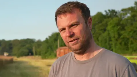 Farmer Olly Harrison with short brown hair and stubble wearing a beige t-shirt. He is standing in a field with trees behind him on a sunny day.