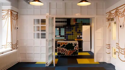 Melanie Issaka A room with black and yellow squares overlaid on a blue floor. The kitchen wall tiles are blue, yellow, black and white with a matching kitchen island. There are white French doors leading into another room with gold curtains painted on the white wall. 