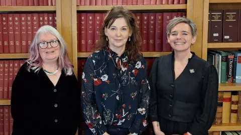 Suzy Angus with the Lord Advocate Dorothy Bain KC and Emma Bryson. They are standing in front of book shelves. Suzy has long white and pink hair and is wearing a black buttoned cardigan. Dorothy Bain wears a navy blue blouse with red and white floral print. Emma has a dark blue blouse.