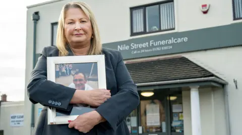 A woman wearing a black suit stands and looks into the camera whilst holding a framed photograph of a man. A building with the words Nile Street Funeralcare is behind her.