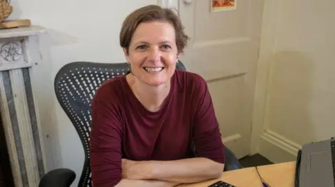 LDRS Helen Bedser from Julian House sits smiling at an office desk. She is wearing a dark red long-sleeved top and is sitting on a modern-style office chair