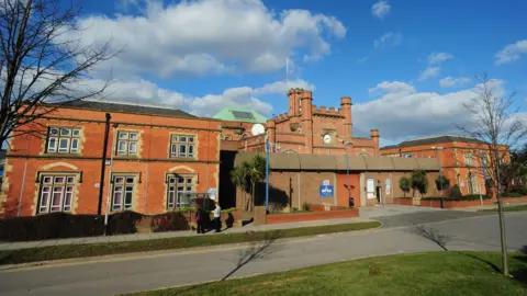 The main gate of HMP Hull on Hedon Road. A red-brick turret and clock tower sits in the centre of the building and stands behind a large concrete wall and red-coloured gate. Two red-brick buildings stand either side of the gate and are surrounded by trees and shrubs. Two people are standing at a bus stop with their backs to the camera.