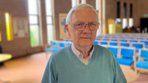 Philip wearing blue v-neck jumper and a checked shirt inside - standing inside the main prayer hall of the church- with blue chairs in the background