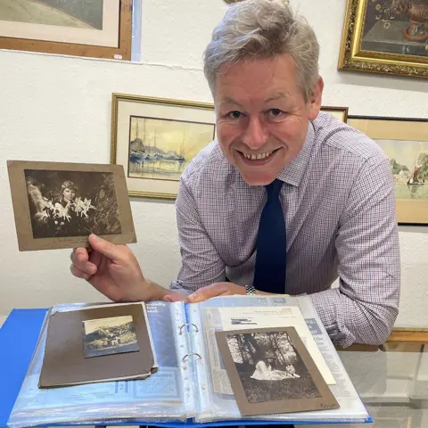 John Taylors Auction Rooms A man with grey hair, a purple and white square patterned shirt and a navy tie is holding a black and white photo of a girl with fairies in front of her. Behind him are paintings on a wall.