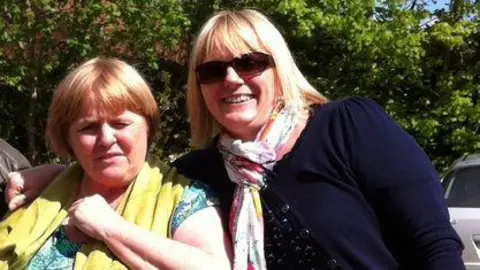 Family handout Shelagh Goodwin and Lindsay Murphy, standing side by side, looking at the camera. Shelagh, on the left is Shelagh, squinting her eyes, wearing a yellow scarf and green top, Lindsay is next to her wearing sunglasses, a multicoloured scarf and blue top and cardigan. They are standing in front of trees. 