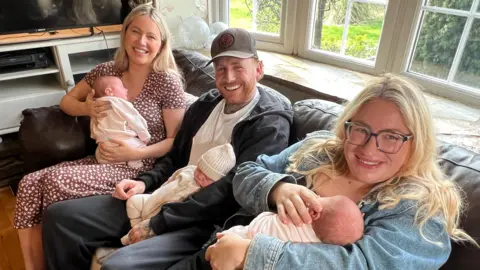 The three parents are sitting on a sofa each holding one of the babies. They are all smiling at the camera.