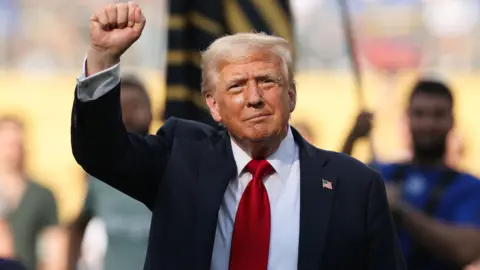 Donald Trump, who has grey/ blonde hair combed to the side, raises his first aloft as he salutes a crowd at a football match in New York. He is wearing a white shirt, red tie and a navy suit. Blurred figures can be seen in the background, including a man holding a flag.