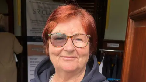 Shows a woman with red hair and glasses smiling. She is wearing a blue hoodie.