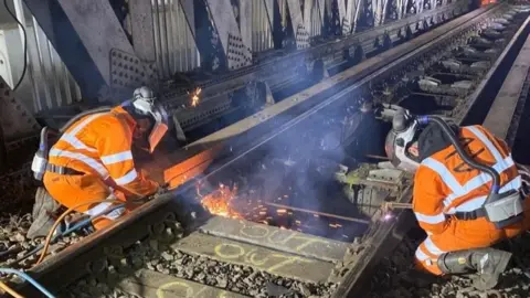 Work taking place on the bridge. Two engineers are welding next to the railway track. They wear high visibility orange jackets and face masks. 